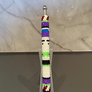 May include: A decorative pen with a colorful beaded design. The pen features a pattern of white, green, black, purple, and red beads. The pen is displayed on a clear acrylic stand.