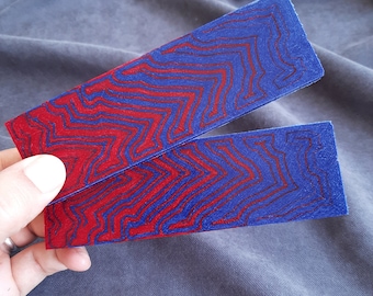 May include: Two rectangular pieces with a blue and red starburst pattern. The pieces are held against a gray fabric background. The red lines are outlined with a darker red color.