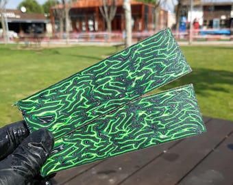 May include: Two rectangular pieces with a green and black abstract pattern are held by a gloved hand. The pattern resembles a network of veins or a topographical map. The pieces are displayed outdoors on a wooden surface.