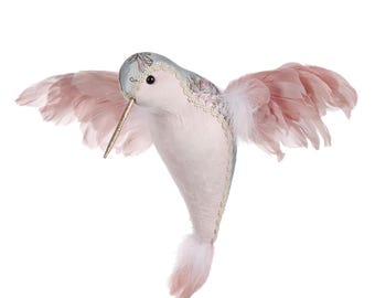 Könnte beinhalten: Ein verspielter, plüschiger Kolibri mit ausgebreiteten Flügeln. Der Vogel hat einen hellblauen Kopf mit floralen Akzenten, einen langen goldenen Schnabel und weiche rosa Federn. Sein Körper ist ein zartes Rosa, und die Flügel sind ein zartes Rosa. Der Kolibri ist so gestaltet, als ob er im Flug wäre.