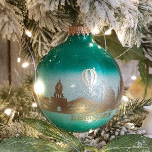 Könnte beinhalten: Ein grüner Weihnachtsbaumschmuck mit einem weißen und goldenen Stadtbild. Der Schmuck zeigt einen Heißluftballon und das Wort "Dresden" in goldenen Buchstaben.