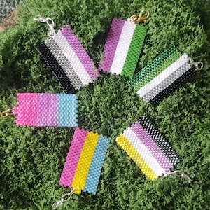May include: A collection of beaded keychains in various colors, arranged on a bed of green moss. The keychains feature the colors of different pride flags, including pink, blue, white, black, and yellow. Each keychain has a silver or gold clasp.