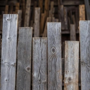 Op de afbeelding: Een verweerde houten schutting gemaakt van verticale planken. De planken zijn in verschillende tinten grijs en bruin, met zichtbare houtnerf en knopen. De achtergrond bestaat uit meer houten planken, wat een rustieke, getextureerde achtergrond creëert.