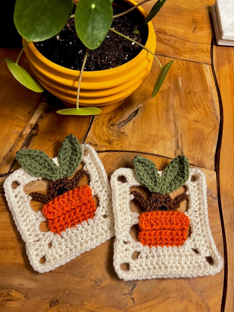 May include: Two crocheted squares with a plant design. Each square features a brown pot with an orange plant and green leaves. The squares are white with a cream color.
