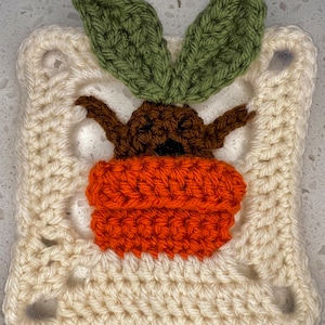 May include: A crocheted square featuring a brown and orange carrot with green leaves. The square is made with white yarn and has a textured, crocheted design.