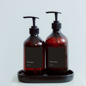 Könnte beinhalten: Zwei braune Kunststoff-Shampoo- und Duschgelflaschen mit schwarzen Pumpen und schwarzen Etiketten, die auf einem schwarzen Tablett stehen. Die Shampooflasche ist mit "Shampoo" beschriftet. Die Duschgelflasche ist mit "Body Wash" beschriftet.