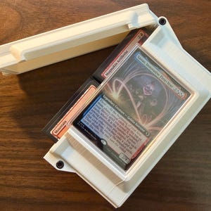 May include: A white 3D-printed plastic card storage box with a hinged lid, containing a stack of trading cards. The top card is a Sanar, Innovative First-Year legendary creature card, with a Magmakin Artillerist card and a Mountain card visible.