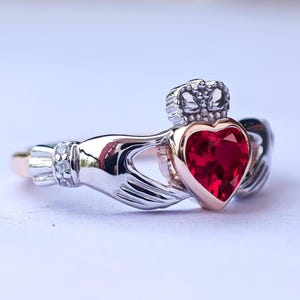 May include: A silver and rose gold Claddagh ring with a red heart-shaped gemstone. The ring features two hands clasping the heart, symbolising love, loyalty, and friendship.