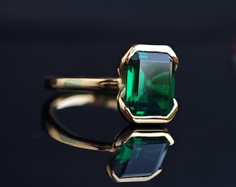 Op de afbeelding: Een gouden ring met een grote, rechthoekige, smaragdgroene edelsteen. De edelsteen is gezet in een gouden bezel, en de ringband is ook goud. De ring wordt gepresenteerd op een reflecterend oppervlak.