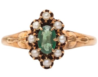 May include: A gold ring featuring a central oval-cut green gemstone surrounded by a cluster of eight small white pearls. The metal band displays a smooth, polished finish with decorative leaf-like engravings on the sides of the setting.