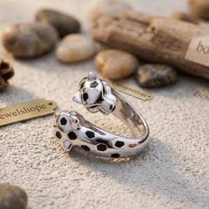 Könnte beinhalten: Ein silberner Ring mit Leopardenmuster. Der Ring hat an jedem Ende einen Leopardenkopf mit schwarzen Flecken und weißen Akzenten. Der Ring liegt auf einer strukturierten Oberfläche mit kleinen Steinen und Holz im Hintergrund.