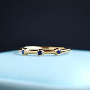 Può includere: Un delicato anello d'oro con tre piccole gemme rotonde blu scuro. L'anello ha una fascia sottile e un design minimalista, perfetto per l'uso quotidiano. Le gemme sono uniformemente distanziate lungo la parte superiore dell'anello.