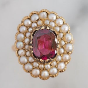 May include: A gold ring featuring a central oval-cut deep pink gemstone held by prongs. The stone is surrounded by a double halo of small, round, cream-colored pearls set in a scalloped gold frame, creating a floral-like cluster design.