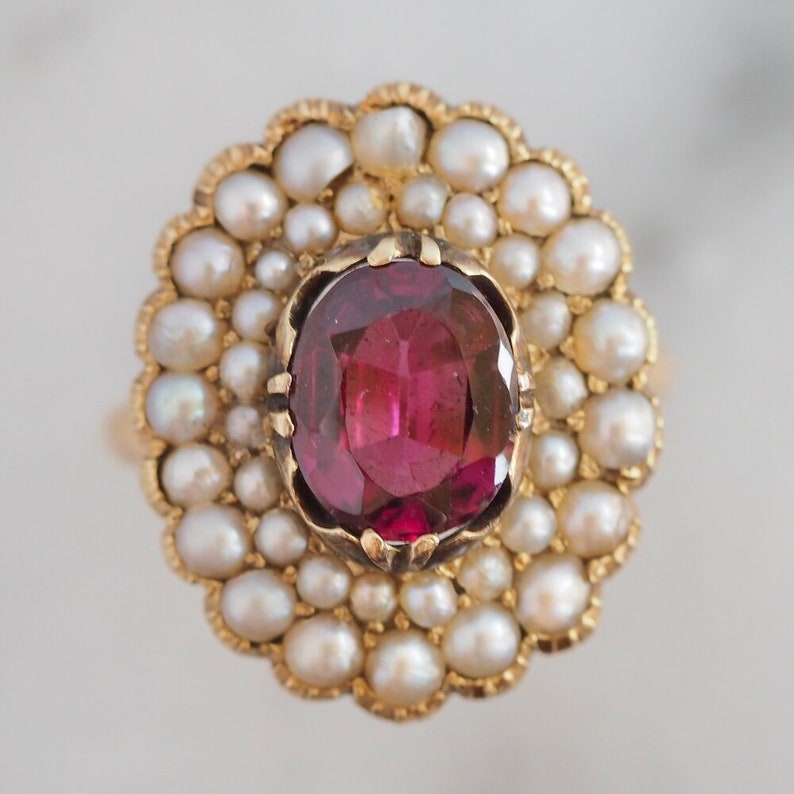 May include: A gold ring featuring a central oval-cut deep pink gemstone held by prongs. The stone is surrounded by a double halo of small, round, cream-colored pearls set in a scalloped gold frame, creating a floral-like cluster design.