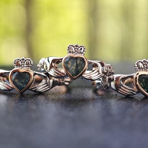 May include: Three silver and gold Claddagh rings with green stone hearts. The rings are stacked on top of each other.