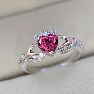 May include: A silver Claddagh ring featuring a heart-shaped, vibrant pink gemstone. The heart is held by two hands, with a crown above the heart. The ring is a symbol of love, loyalty, and friendship.