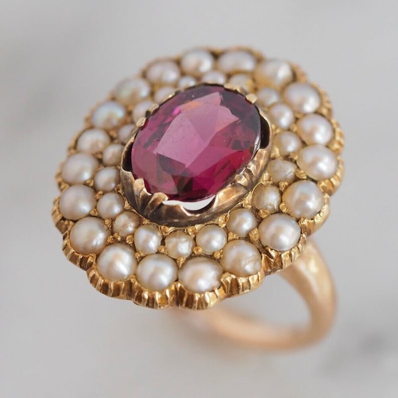 May include: A gold ring featuring a large, oval-cut, deep pink gemstone at the center, surrounded by a double halo of small, round, white pearls set in a scalloped gold frame. The band is plain gold, and the setting has a textured finish.