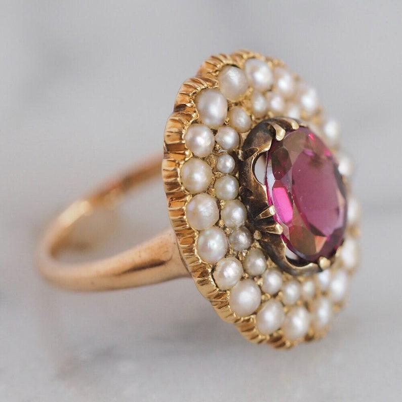 May include: A gold ring featuring a central oval-cut deep pink gemstone surrounded by a double halo of small white seed pearls. The setting has a scalloped edge, and the band is a smooth, polished gold metal, creating a circular cluster design.