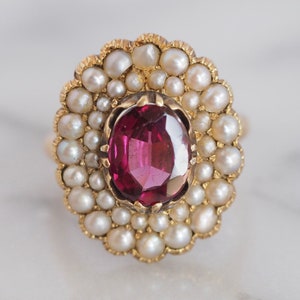 May include: A gold ring featuring a central oval-cut deep pink gemstone surrounded by a halo of small, round, cream-colored pearls. The setting has a scalloped edge, and the stones are mounted in a yellow gold metal band with a polished finish.