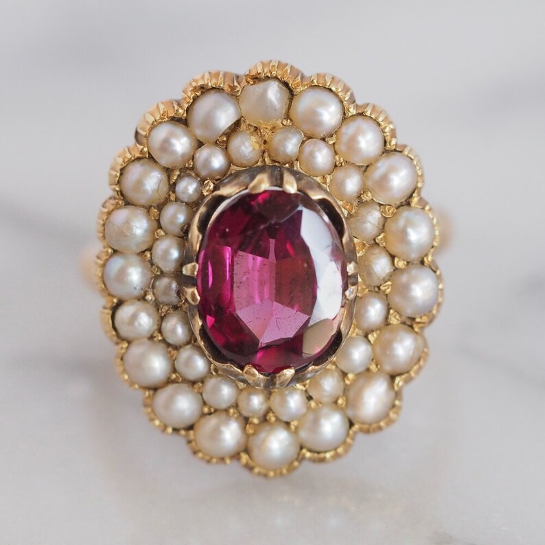 May include: A gold ring featuring a central oval-cut deep pink gemstone surrounded by a halo of small, round, cream-colored pearls. The setting has a scalloped edge, and the stones are mounted in a yellow gold metal band with a polished finish.