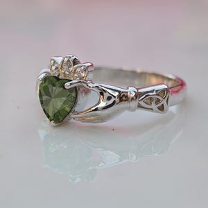 May include: A silver Claddagh ring with a heart-shaped green gemstone. The ring features a crown and hands holding the heart, with a Celtic knot design on the band. The ring is a symbol of love, loyalty, and friendship.