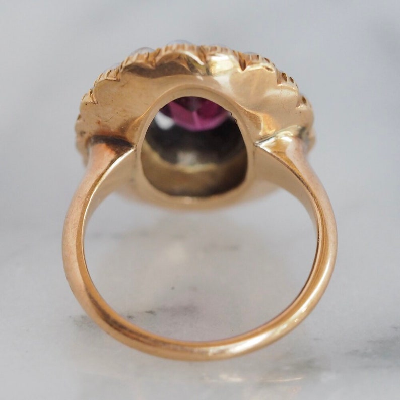 May include: A gold ring featuring a central oval-shaped deep pink gemstone surrounded by a scalloped border of small white pearls. The band is smooth and circular, showcasing the back view of the setting with a polished metallic finish.