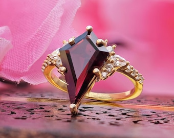 May include: A gold ring with a large, dark red, faceted gemstone in a unique, pointed shape. The ring is set with smaller, clear gemstones around the main stone.