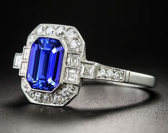 May include: An Art Deco-style ring featuring a large, rectangular, deep blue gemstone as the focal point. The gemstone is surrounded by smaller, clear gemstones in a geometric pattern, set in a silver-colored band. The ring is displayed against a dark background.