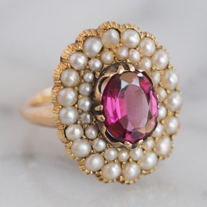 May include: A gold ring featuring a central oval-cut deep pink gemstone surrounded by a halo of small white seed pearls. The setting has a scalloped edge, and the band is made of polished yellow gold, creating a floral-inspired cluster design.
