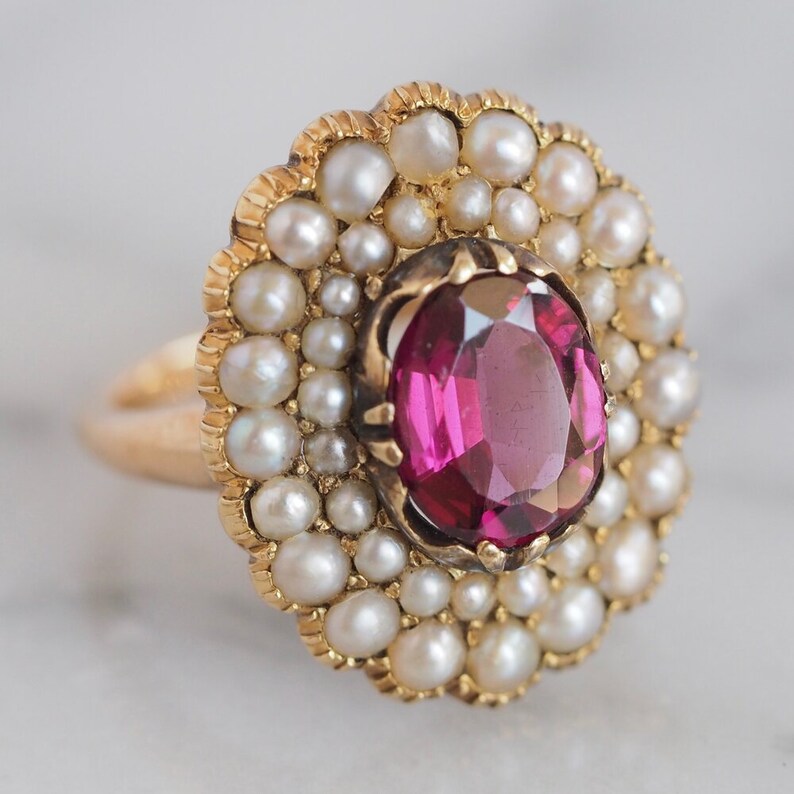 May include: A gold ring featuring a central oval-cut deep pink gemstone surrounded by a halo of small white seed pearls. The setting has a scalloped edge, and the band is made of polished yellow gold, creating a floral-inspired cluster design.