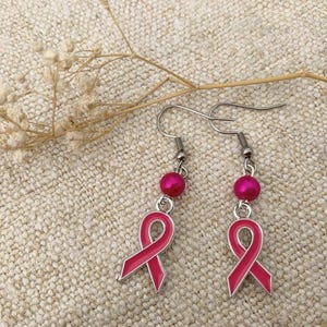 May include: A pair of silver earrings with pink ribbon charms and pink pearl accents. The earrings are on a light brown fabric background.