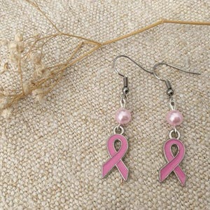 May include: A pair of pink ribbon earrings with silver hooks and pink pearl beads. The earrings are on a light brown fabric background.