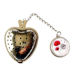 Puede incluir: Un infusor de té de corazón plateado con una cadena y un encanto redondo de vidrio con una flor de amapola roja y un diente de león.