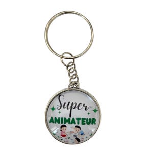 Könnte beinhalten: Ein silberner Schlüsselanhänger mit einem runden, weißen Glasanhänger. Der Anhänger hat grünen Glitzer und den Text "Super Animateur" in Schwarz. Auf dem Anhänger sind drei spielende Comic-Kinder zu sehen.