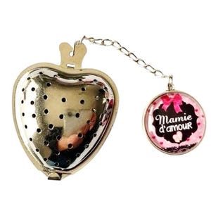 Puede incluir: Un infusor de té con forma de corazón con un acabado plateado y una cadena unida a un charm redondo rosa y negro con el texto "Mamie d'amour".