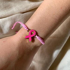 Op de afbeelding: Roze lint bedelarmband. De armband heeft een felroze lintbedel, een symbool voor borstkankerbewustzijn. De armband is gemaakt van roze gevlochten koord en wordt om de pols gedragen.