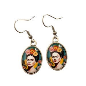 Puede incluir: Un par de pendientes colgantes plateados. Cada pendiente presenta un colgante ovalado con la imagen de una mujer con flores en el pelo. La mujer tiene el pelo oscuro y una expresión seria. El fondo es turquesa y amarillo.
