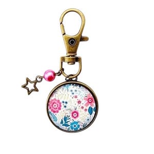 May include: A bronze key chain with a round glass charm featuring a floral pattern in pink, blue, and white. The charm is attached to a star and a pink bead.