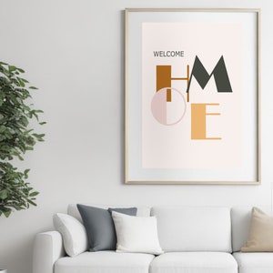 May include: A framed print with a pink background and the word "WELCOME" in black text above the word "HOME" in stylized, colourful letters. The letters are in various shades of brown, green, and pink.