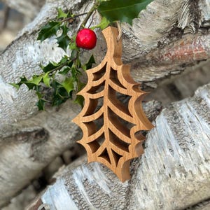 May include: A wooden Christmas ornament in the shape of a holly leaf, with cut-out details. The ornament is a light brown color and hangs from a natural twine. A sprig of holly with green leaves and a red berry is in the background.