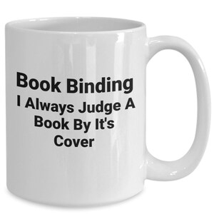 May include: White ceramic coffee mug with black text that reads "Book Binding I Always Judge A Book By It's Cover"