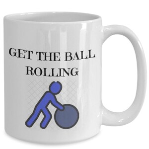 May include: White ceramic mug with the text "GET THE BALL ROLLING" in black. A blue stick figure is pushing a blue ball. The mug has a curved handle and is suitable for hot or cold drinks.