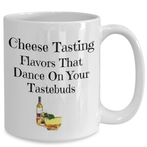 May include: White ceramic coffee mug with a black graphic that says "Cheese Tasting Flavors That Dance On Your Tastebuds". The graphic also includes a bottle of wine, grapes, cheese, and nuts.