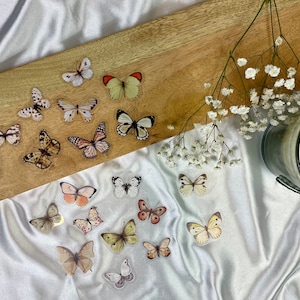 May include: A wooden tray with a gold handle, decorated with butterfly stickers. The tray is on a white fabric surface with a white candle in a gray glass jar and white flowers.