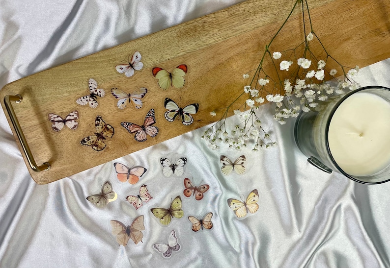 May include: A wooden tray with a gold handle, decorated with butterfly stickers. The tray is on a white fabric surface with a white candle in a gray glass jar and white flowers.