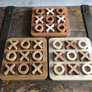 Può includere: Tre giochi di tris in legno con pezzi di diversi colori. Il gioco in alto ha cerchi marroni e X bianche. Il gioco al centro ha cerchi marroni e X marroni. Il gioco in basso ha cerchi bianchi e X marroni. Il gioco in alto ha il testo "Familie Löhn" inciso sulla tavola.