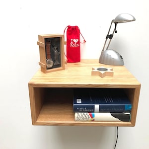 May include: A light wood floating nightstand with a lower shelf. On top, a small lamp, a red bag with "I Love Köln" printed on it, a wooden box with a watch and Statue of Liberty, and a star-shaped candle holder. Two books are on the shelf.