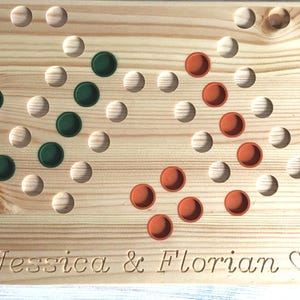 Könnte beinhalten: Holzbrettspiel mit heller Holzmaserung. Das Spiel hat Holzstifte und farbige Spielsteine in Grün, Orange und Gelb. Die Namen "Jessica & Florian" sind in das Holz geschnitzt.
