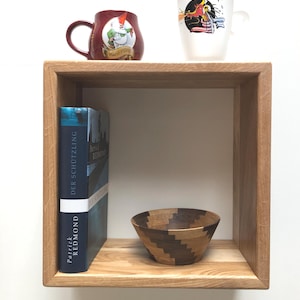 Può includere: Una mensola cubica in legno espone una tazza bordeaux con un gnomo dei cartoni animati, una tazza bianca con un'illustrazione di drago, un libro intitolato "Der Schützling" e una ciotola di legno con un design geometrico.