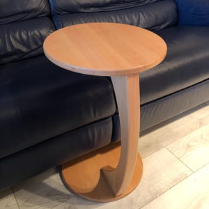 Pode incluir: Uma mesa de apoio de madeira clara com tampo redondo e base curva. A mesa está ao lado de um sofá de couro azul escuro.